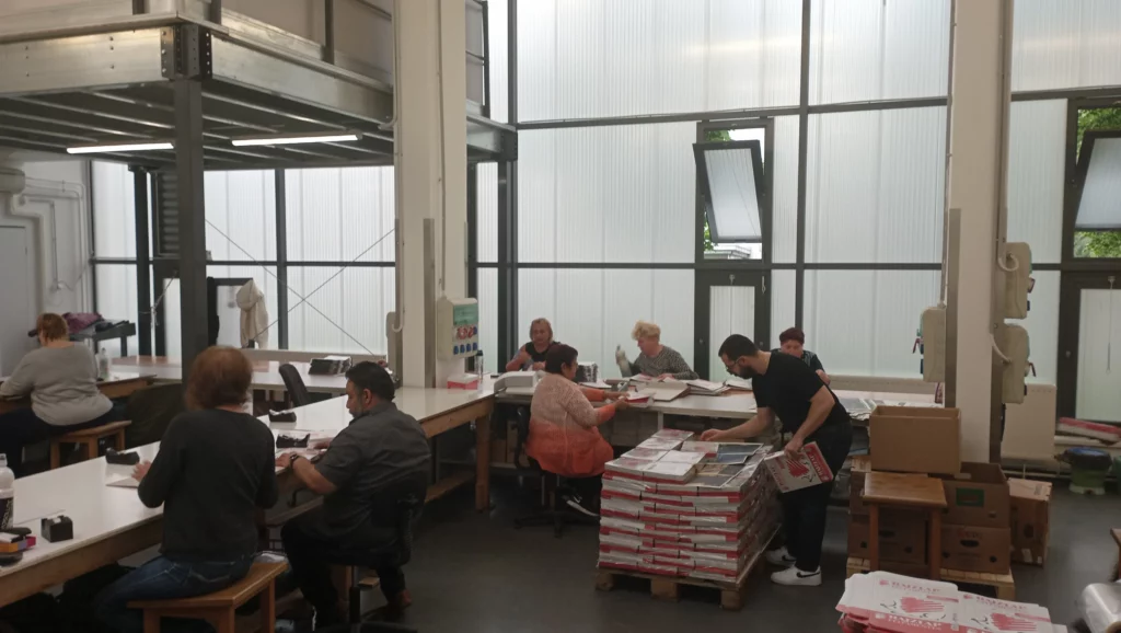 A dolgozók a papíráruk, hajtogatását, rendezését és csomagolását végzik munkaasztalok mellett a csomagoló üzemben.