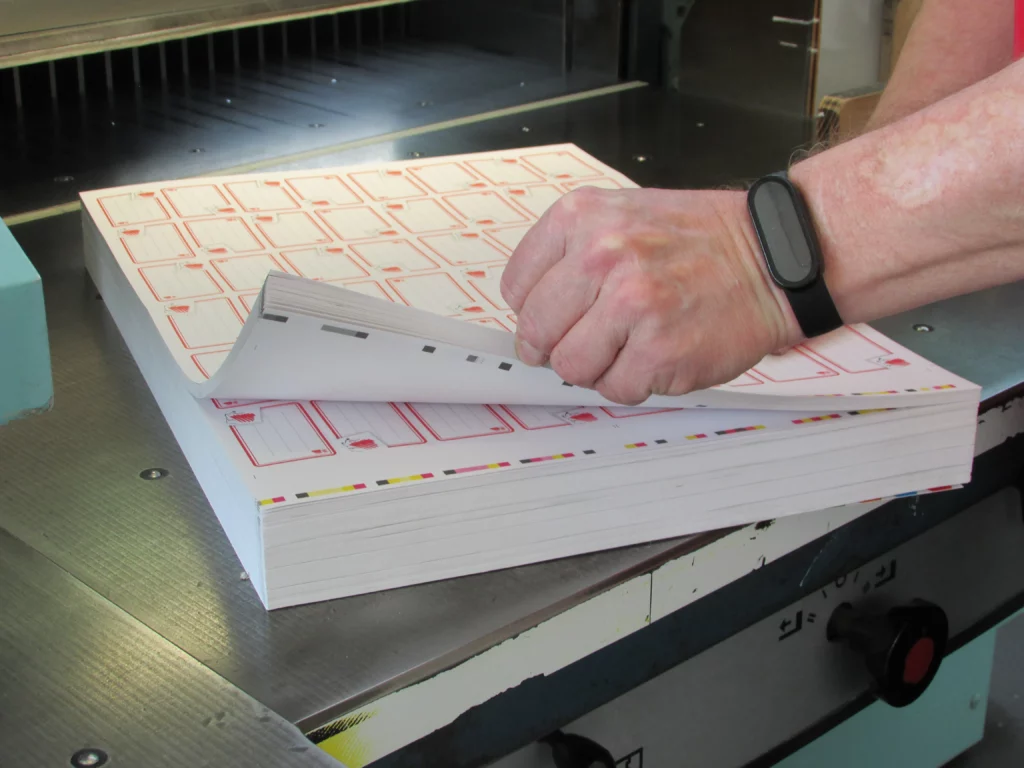 Kötészeti dolgozó öntapadós füzetcímkéket készít elő, hogy a papírvágó gép a riccelési eljárás által azok felső nyomott rétegét átvágja a bemetszési vonalak mentén.